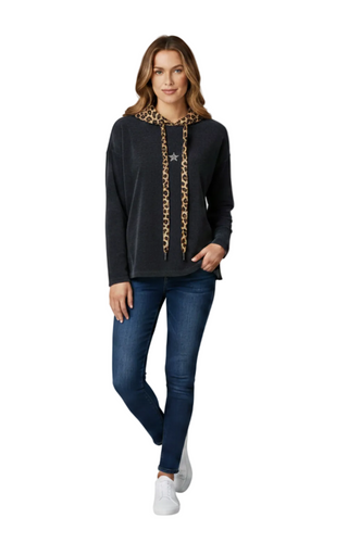 A woman modeling a black long-sleeve top with a gold chain detail down the front, paired with blue jeans and white sneakers, against a plain white background.
