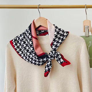 Neck scarf with houndstooth pattern draped over a beige sweater on a wooden hanger against a white background.